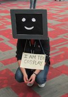 Costume is a computer terminal with smiley face for head; person wears sign saying “I am teh cosplay”